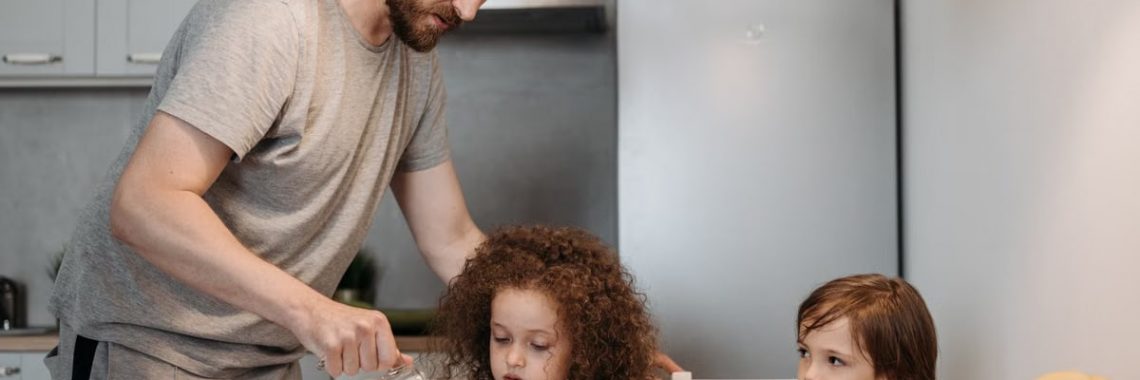 A father pours milk into cereal bowls for his children at the breakfast table in a modern kitchen.