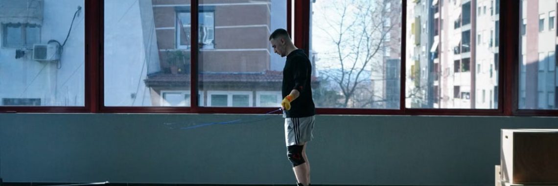 Young man exercising with a skipping rope in an urban gym with large windows and city view.